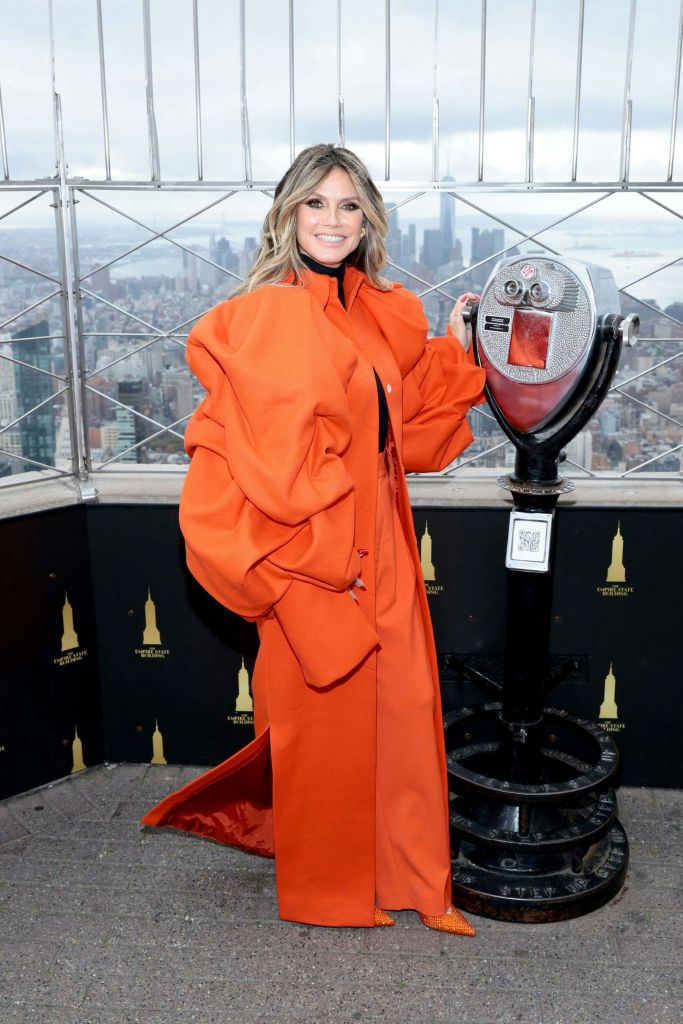 주황색 의상을 입은 사람이 도시 스카이라인이 있는 전망대에서 포즈를 취하고 있으며, 배경에는 양안시경이 있습니다.