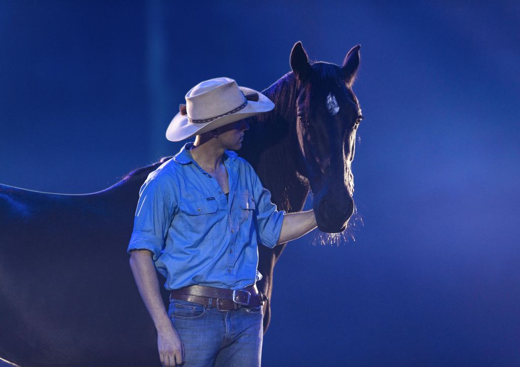 파란색 셔츠와 카우보이 모자를 쓴 사람이 파란색 조명이 켜진 어두운 환경에서 다크호스 옆에 서 있습니다.