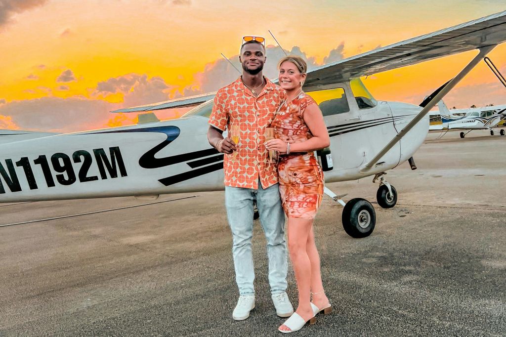 해가 질 무렵 주황색 의상을 입은 커플이 작은 비행기 앞에 서서 음료를 들고 미소를 짓고 있습니다.