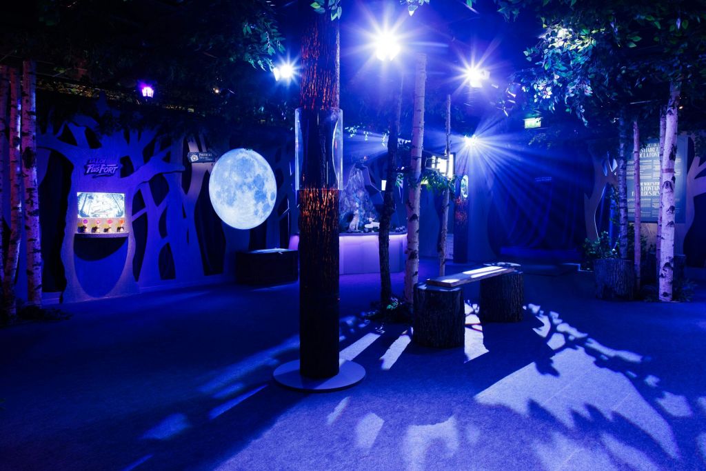 인공 나무, 커다란 달 배경, 파란색 조명, 나무 그루터기 좌석이 있는 희미한 조명의 실내 숲 테마 공간입니다.