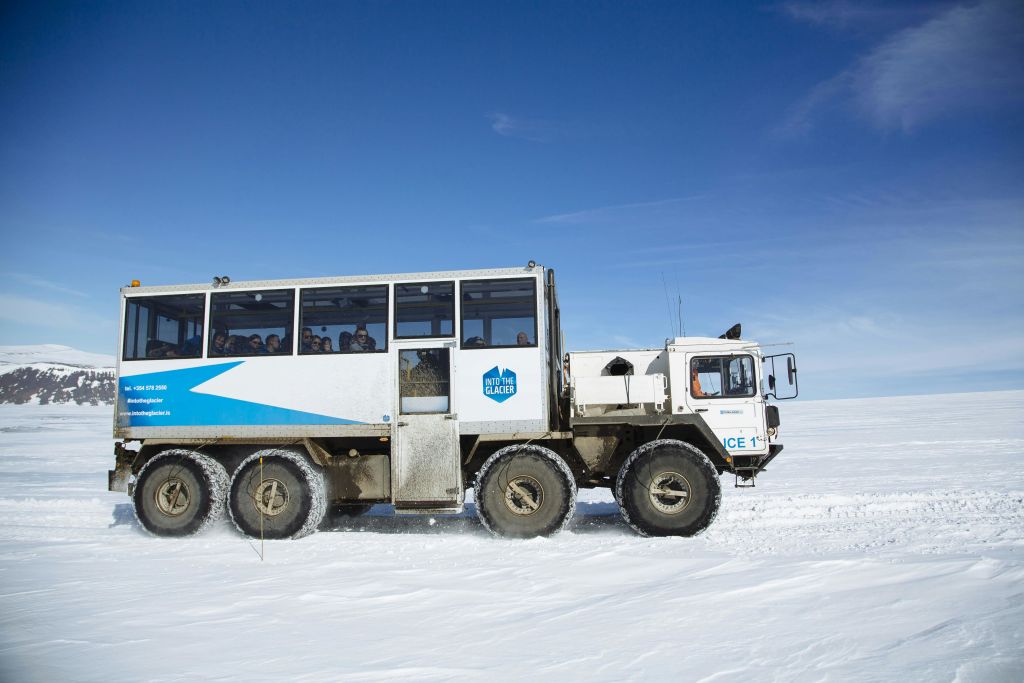 파란 줄무늬와 "Into the Glacier" 로고가 있는 흰색의 대형 오프로드 차량이 맑고 푸른 하늘 아래 눈 덮인 지형을 가로지르고 있습니다.