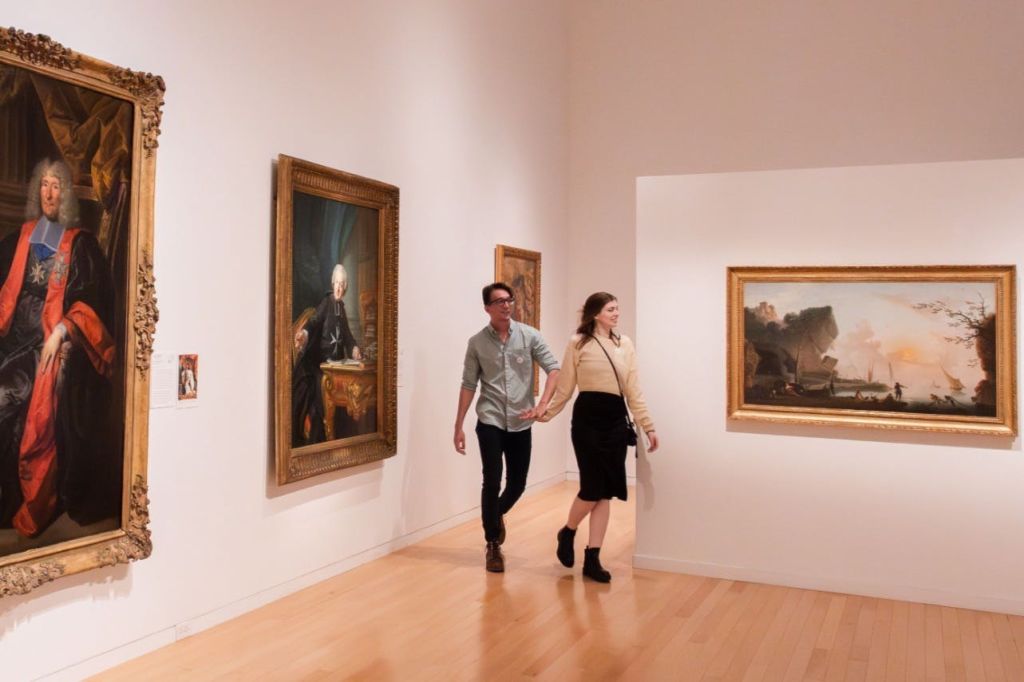 흰색 벽과 나무 바닥에 액자 그림이 놓인 미술관을 지나가는 커플의 모습입니다.