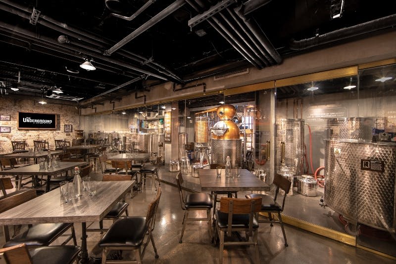 나무 테이블과 의자가 있는 좌석 공간에서 유리를 통해 볼 수 있는 인더스트리얼 스타일의 증류소. 구리 스틸과 강철 탱크가 보입니다.