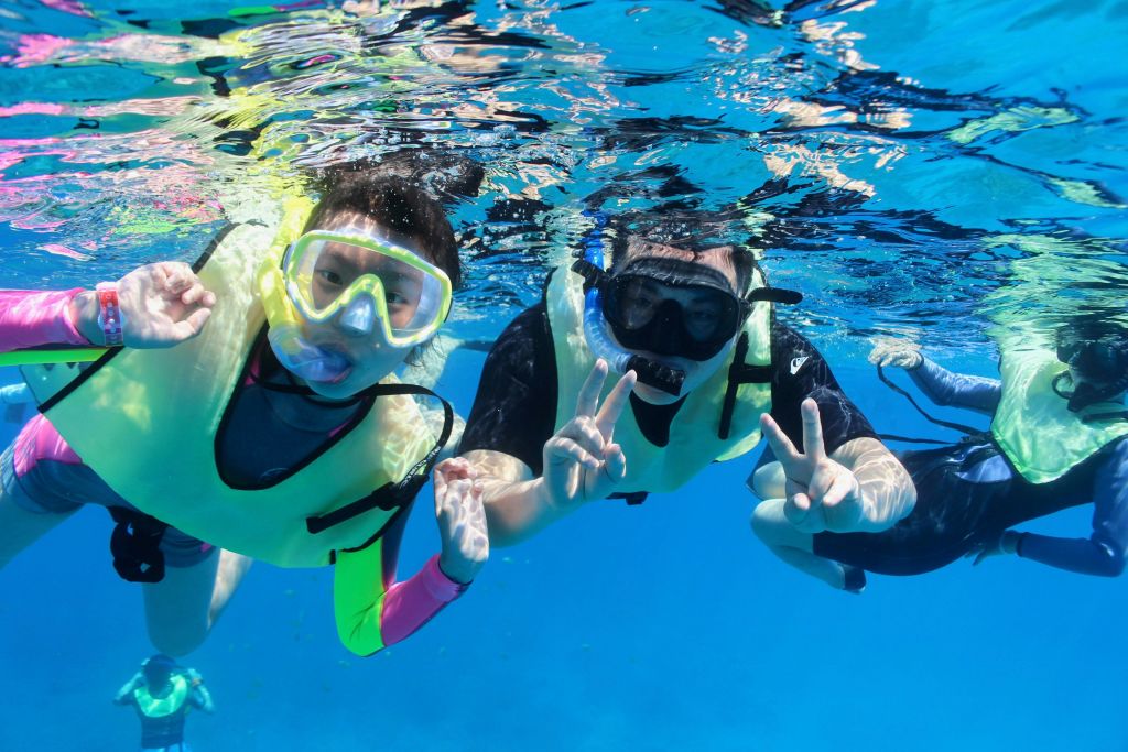 수중에서 스노클링 장비와 구명 조끼를 착용한 두 사람이 맑고 푸른 물 속에서 카메라를 향해 평화의 표시를 하고 있습니다.