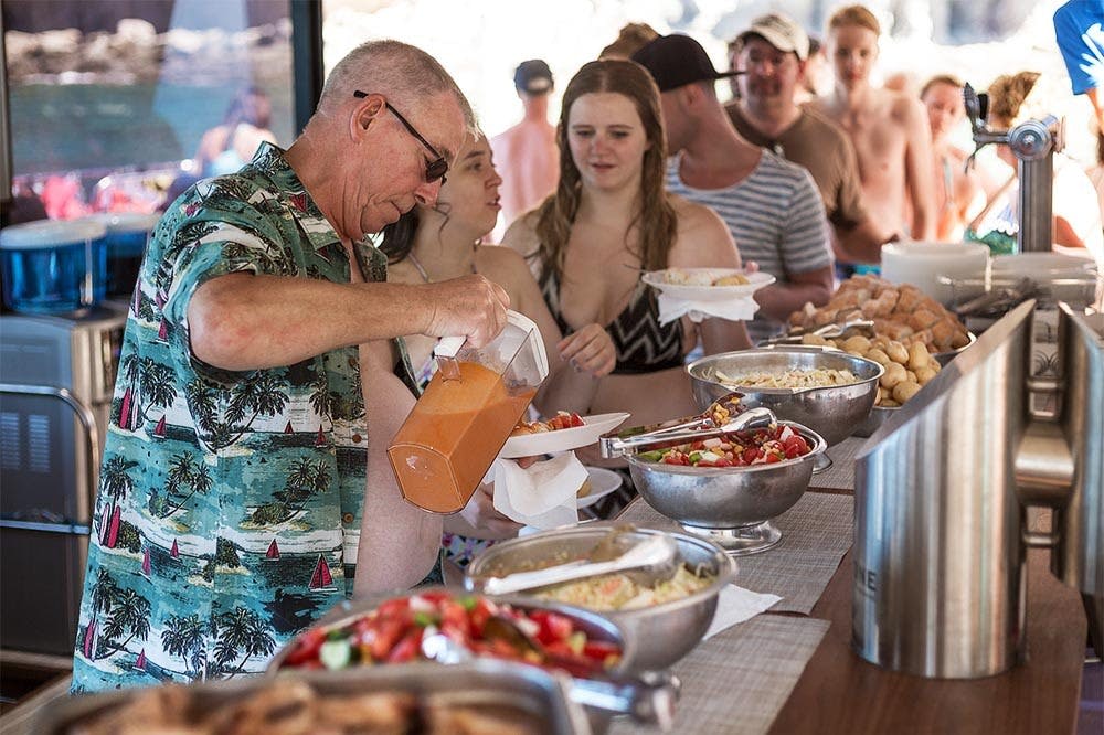 수영복을 입은 한 무리의 사람들이 다양한 요리와 음료 디스펜서가 있는 뷔페에서 음식을 제공하고 있습니다.