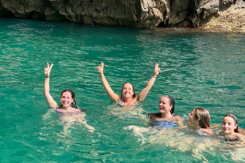 수영복을 입은 세 사람이 바위 절벽 근처의 맑은 청록색 물에서 수영을 즐기며 팔을 들어 올리고 있습니다.