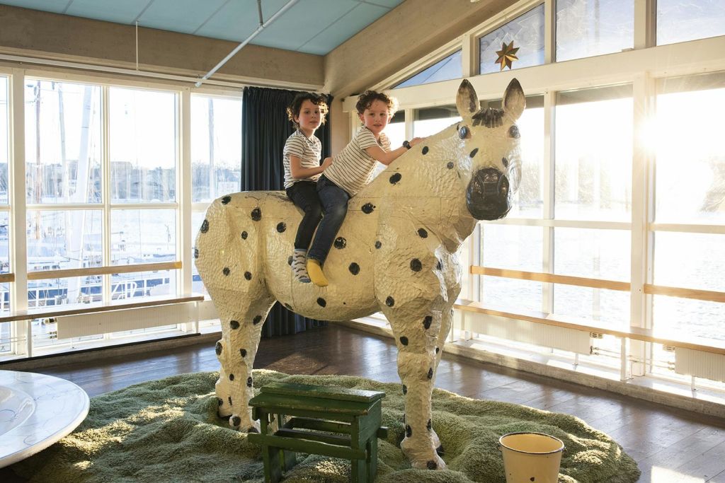 반짝이는 줄무늬 셔츠를 입은 두 어린이가 햇살이 비치는 방 안에 있는 흰색의 커다란 점박이 말 조각상 위에 앉아 있습니다.