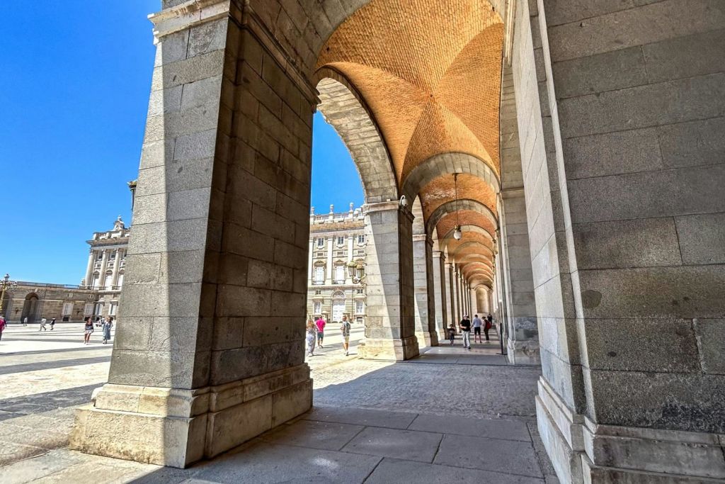 천장이 아치형인 석조 아치형 통로와 그 아래를 걷고 있는 사람들, 역사적인 건물이 배경으로 보입니다.