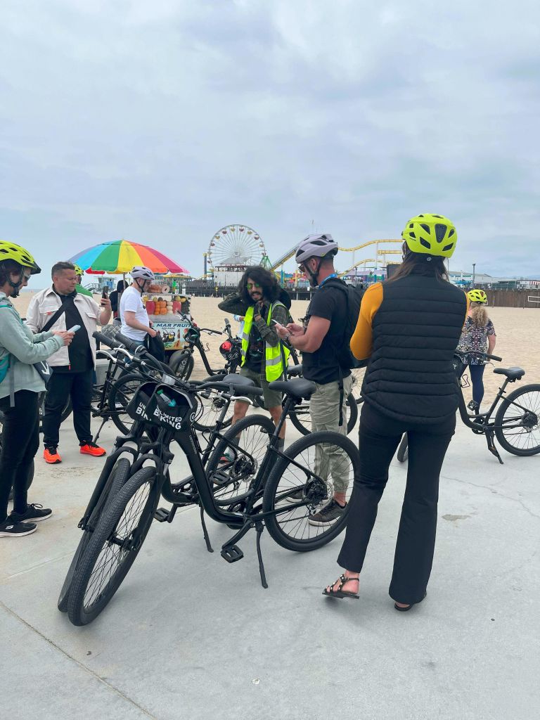 네온색 헬멧과 자전거를 착용한 한 무리의 사람들이 해변의 화려한 우산을 들고 대관람차를 배경으로 상인 가까이 모여듭니다.