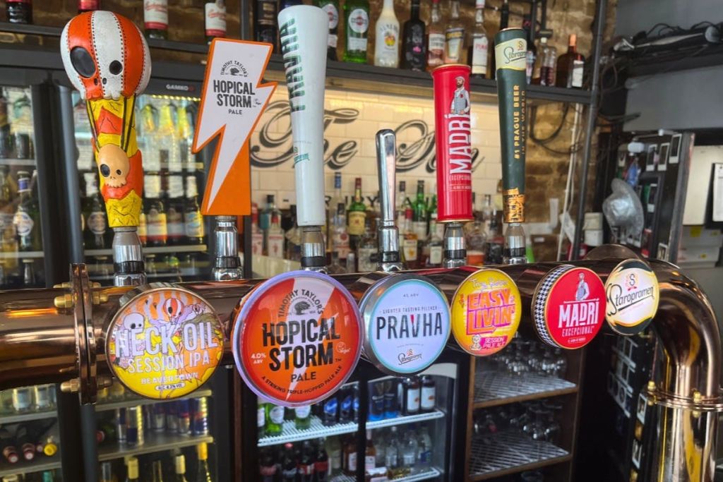 다양한 맥주 브랜드로 레이블이 지정된 바에서 맥주 한 줄을 탭합니다. 배경에는 병이 있는 선반이 보입니다.