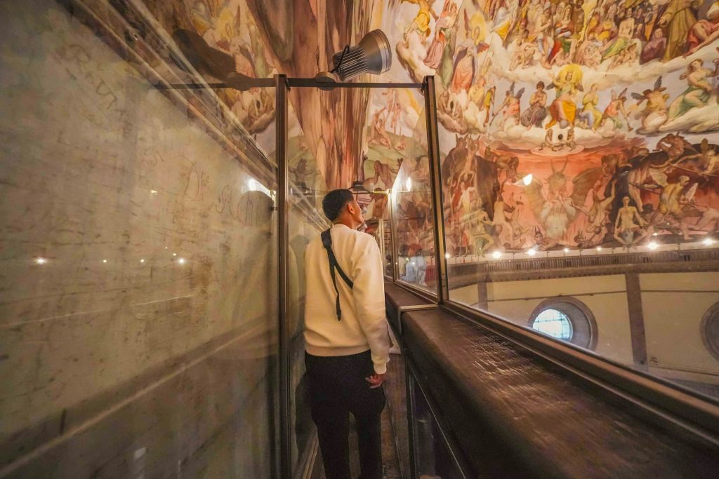 벽과 천장에 화려한 프레스코화로 둘러싸인 유리 장벽이 있는 좁고 높은 통로를 따라 걷는 사람.