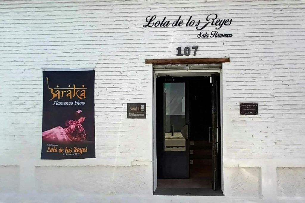 위에 "Lola de los Reyes Sala Flamenca" 라고 쓰여진 하얀 벽돌 건물 입구, 왼쪽에 포스터, 문 위의 숫자 107.
