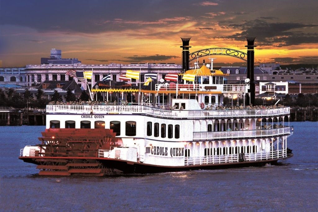 도시를 배경으로 여러 개의 데크와 화려한 색상의 국기가 있는 "크리올 여왕" 이라는 이름의 강 보트가 일몰에 항해합니다.