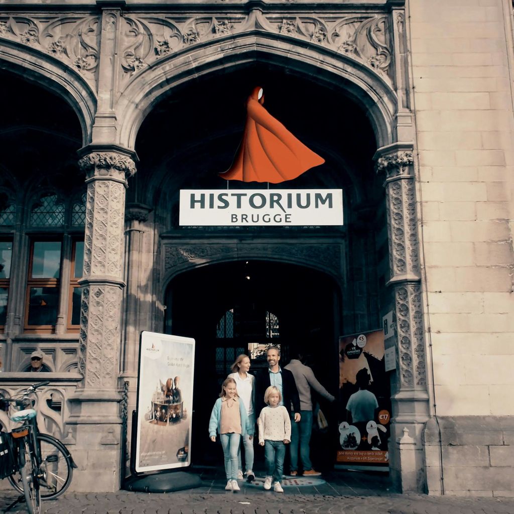 장식용 석조 아치와 간판이 있는 유서 깊은 건물인 Historium Brugge 입구에 4개의 스탠드가 있습니다.