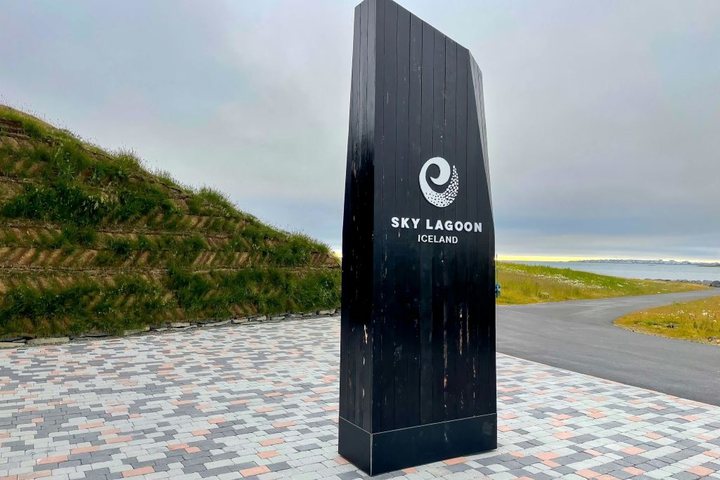 "스카이 라군 아이슬란드 (Sky Lagoon Iceland)" 로고가 새겨진 검은색 간판이 무늬 포장 도로 위에 서 있습니다. 잔디로 뒤덮인 언덕과 흐린 하늘을 배경으로.
