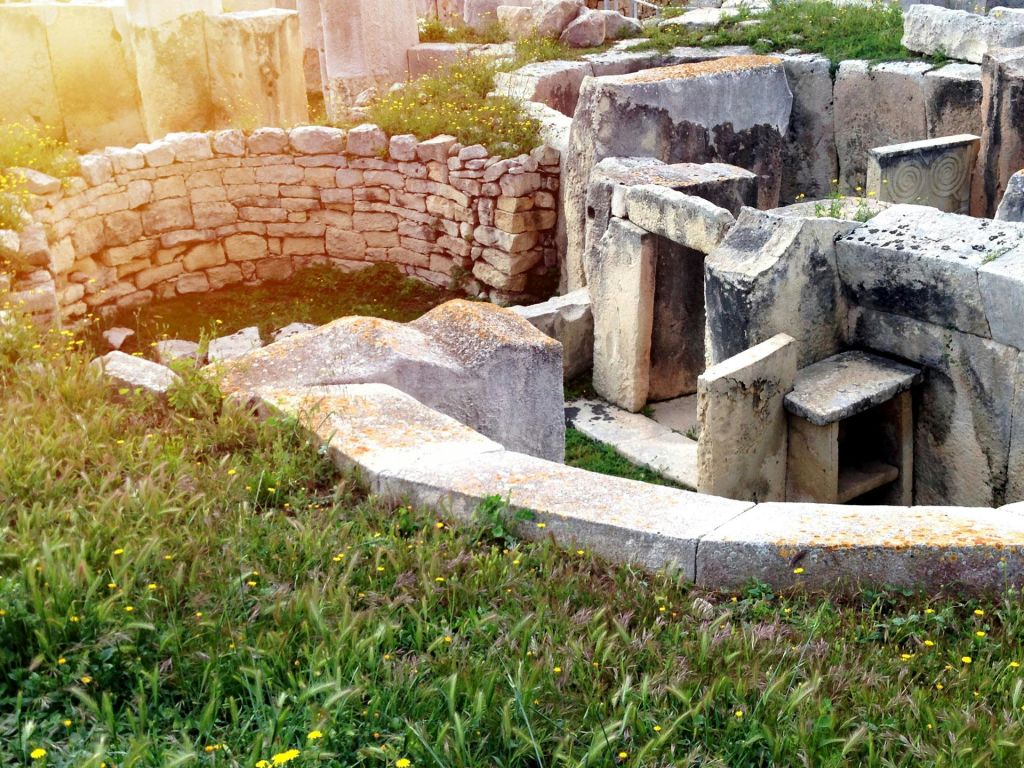 잔디와 작은 노란 꽃으로 둘러싸인 원형 벽과 커다란 석판이 있는 고대 석조 유적지.