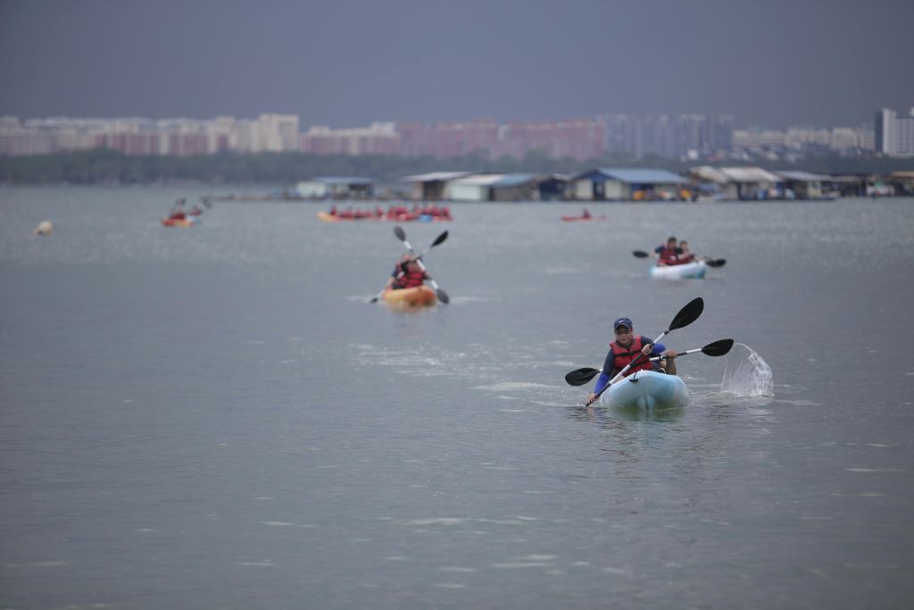잔잔한 물가에서 노를 젓는 카약 타는 사람들, 구름이 잔뜩 낀 하늘 아래 저 먼 배경의 건물과 보트.