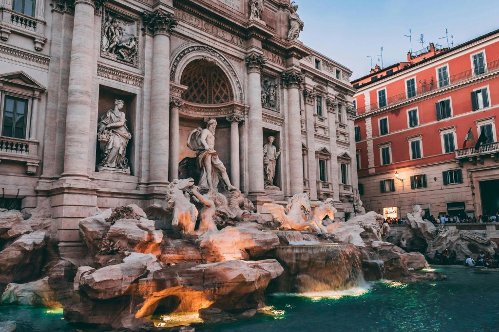 석상과 흐르는 물이 있는 웅장한 바로크 양식의 분수로, 해질 무렵 화려한 건물 외관과 맞닿아 있습니다.