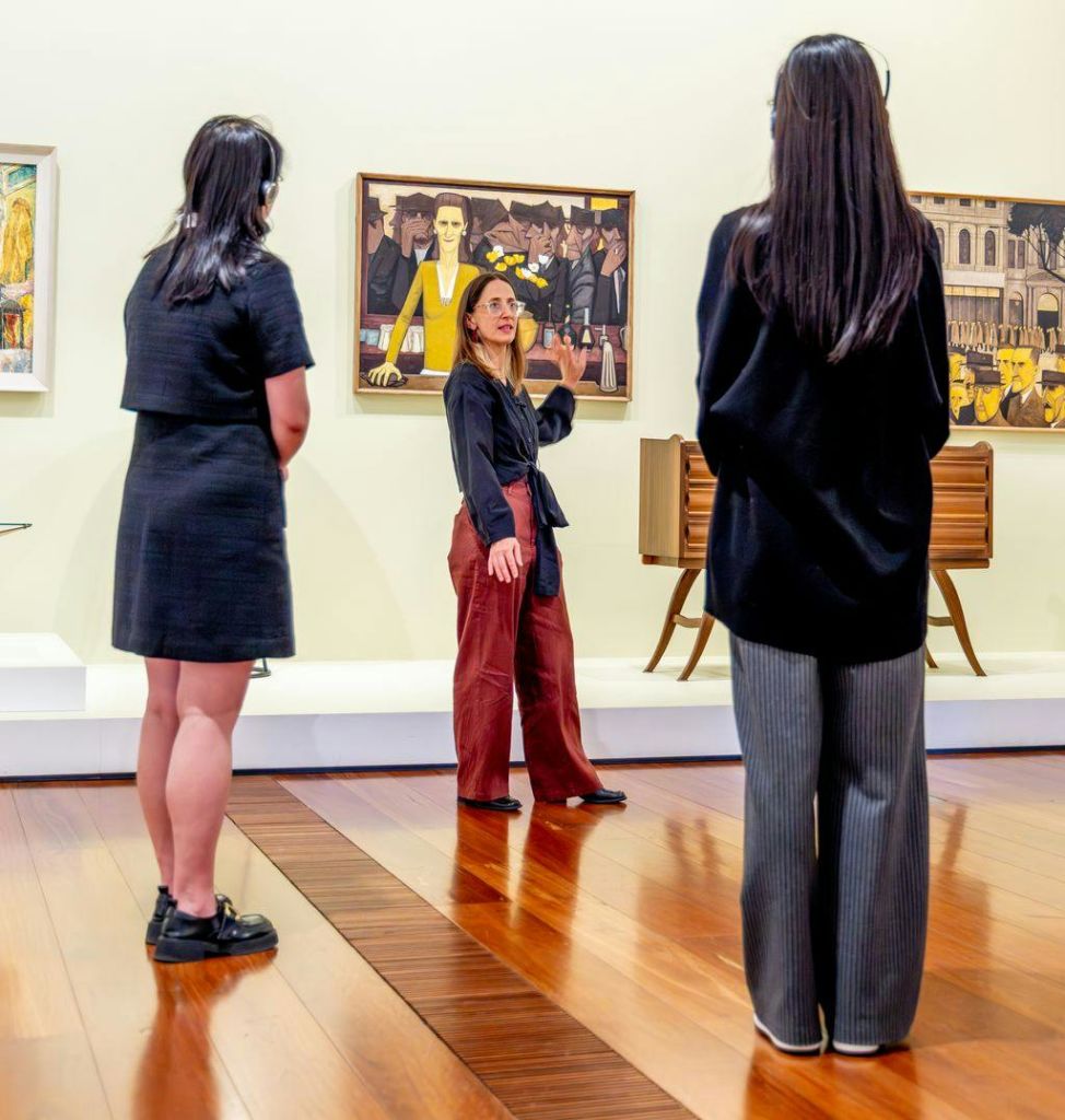 가이드가 벽에 그려진 그림과 광택이 나는 나무 바닥이 있는 갤러리에 있는 두 명의 방문객에게 미술품을 설명합니다.