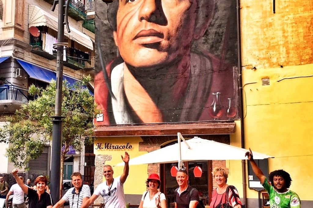 다양한 사람들이 도시 거리에 있는 왕관을 쓴 남자의 커다란 벽화 아래에서 자전거를 들고 포즈를 취하고 있습니다.