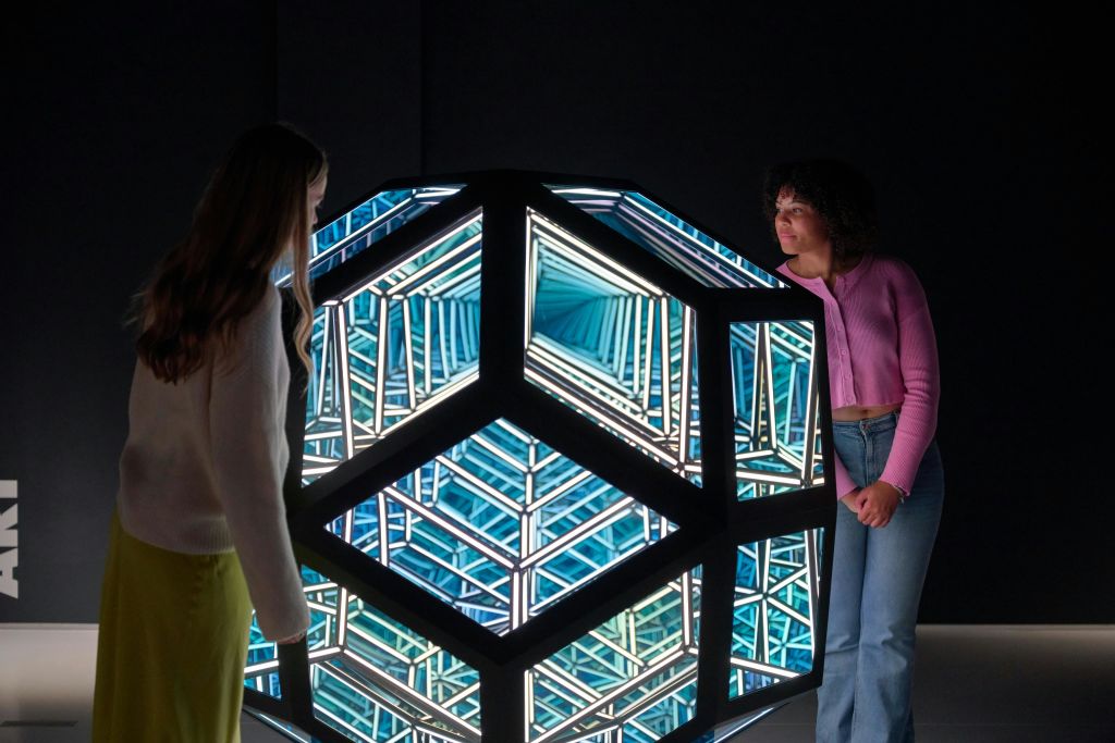 한 사람이 파란색과 흰색 빛을 내는 정육면체 구조의 기하학적 조명 예술 작품 옆에 서 있습니다.