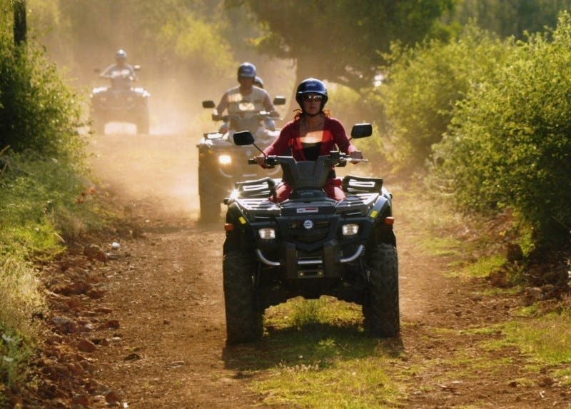 녹지로 둘러싸인 먼지가 쌓인 산책로에서 ATV를 타는 사람들. 승객은 빨간색 의상과 헬멧을 착용합니다.
