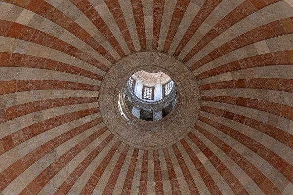 빨간색과 베이지색의 방사형 줄무늬가 번갈아 보이는 돔 천장과 창문과 아치가 있는 구조물이 보이는 오큘러스를 올려다보세요.