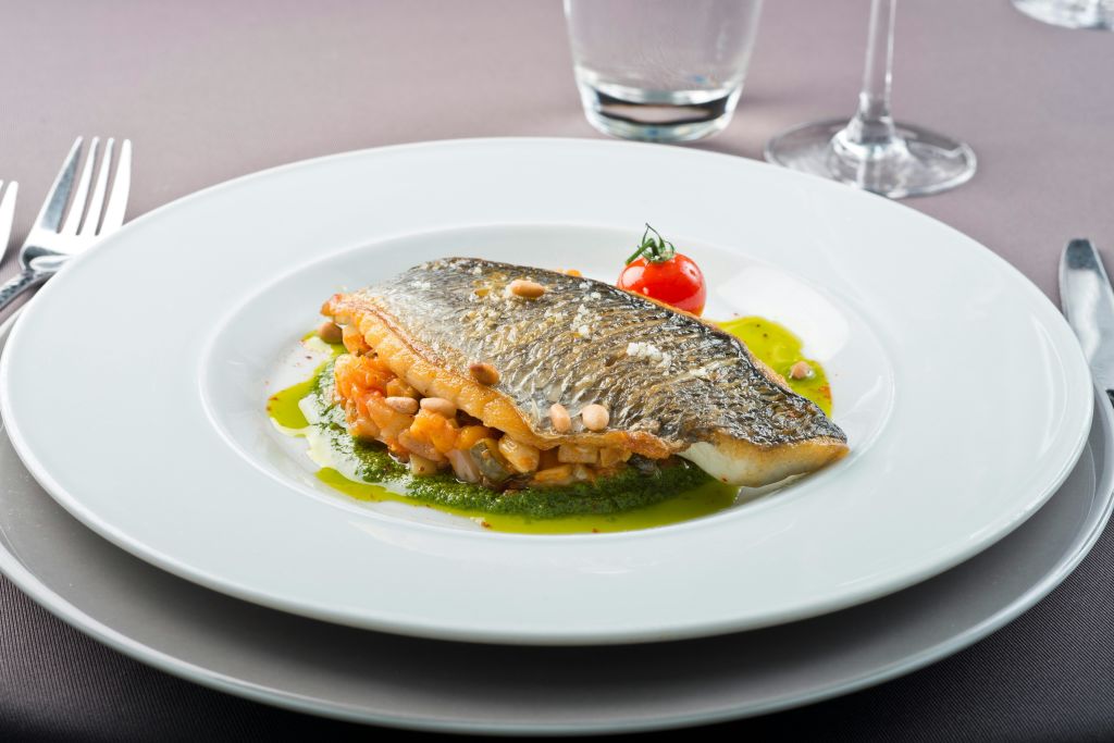 초록색 소스를 곁들인 야채 침대에 조리된 생선 필레가 있는 도금 요리로, 체리 토마토로 장식되어 있습니다.