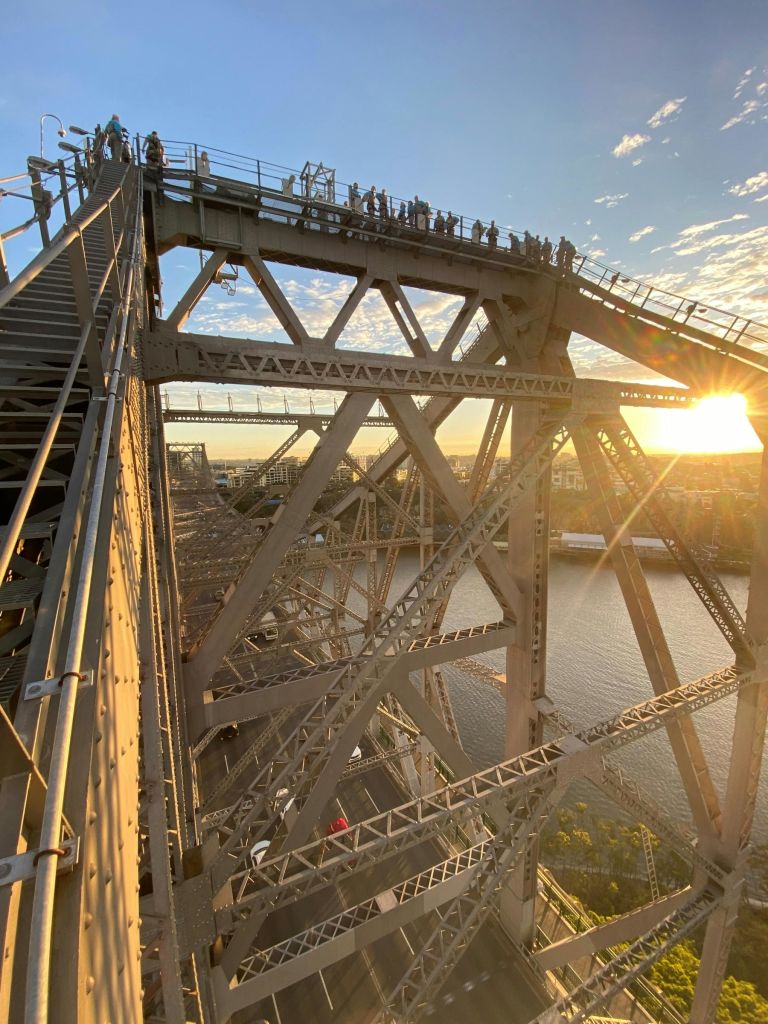 강과 도시 풍경이 내려다보이는 석양에 커다란 철교의 상부 구조물을 걷는 사람들.