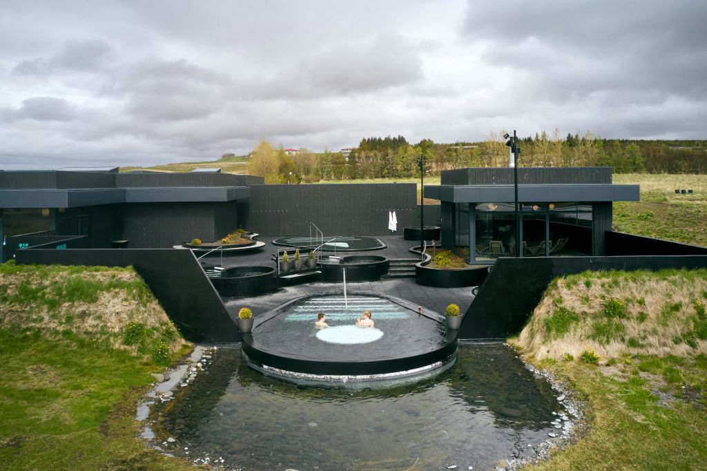 탁한 하늘 아래 어두운 구조와 녹지로 둘러싸인 현대적인 야외 스파에서 두 사람이 원형 온수 욕조에서 휴식을 취하고 있습니다.