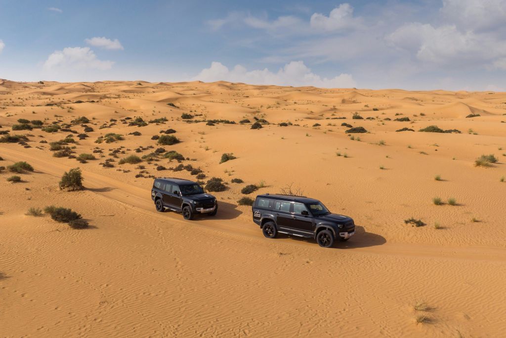 부분적으로 흐린 푸른 하늘 아래 모래 언덕과 희박한 초목이 있는 광활한 사막을 달리는 두 대의 검은 SUV.