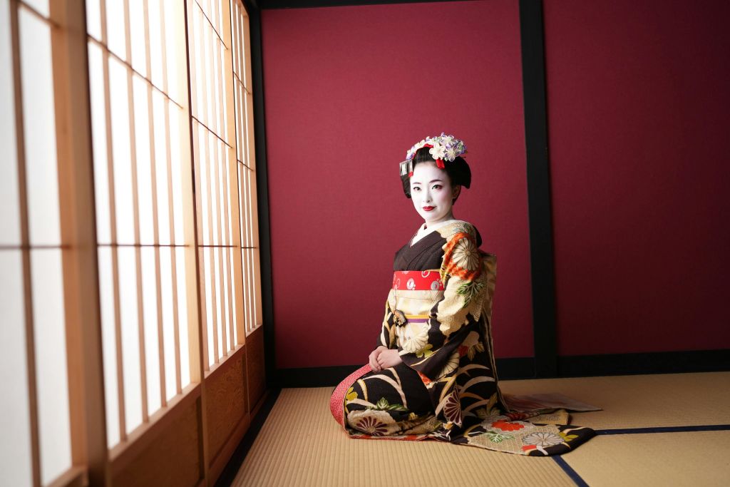 일본 전통 기모노를 입은 여성이 다다미 바닥에 무릎을 꿇고 있으며, 그 옆에는 진분홍색 벽과 쇼지 스크린 창문이 있습니다.