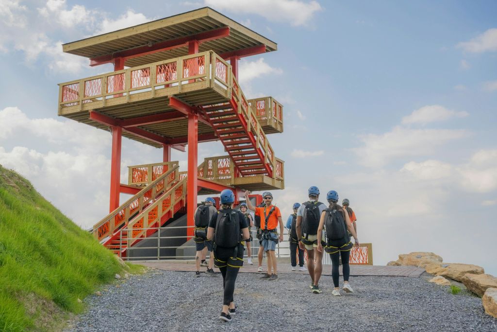 헬멧과 장비를 착용한 한 무리의 사람들이 맑은 하늘 아래 계단이 있는 붉은 나무 전망대에 다가가고 있습니다.