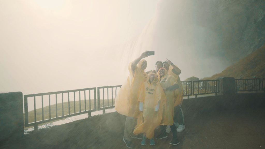 노란색 판초를 입은 한 무리의 사람들이 금속 난간 옆 폭포 앞에서 셀카를 찍고 있습니다.