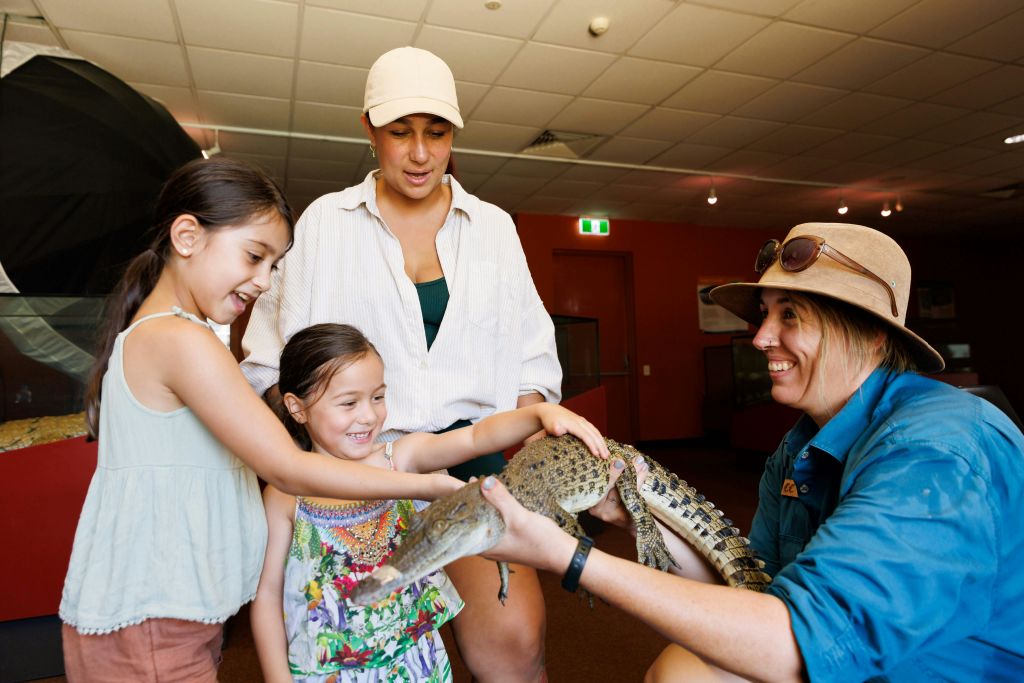 두 아이와 한 여성이 실내에서 동물원이 들고 있는 작은 악어를 쓰다듬고 있습니다.