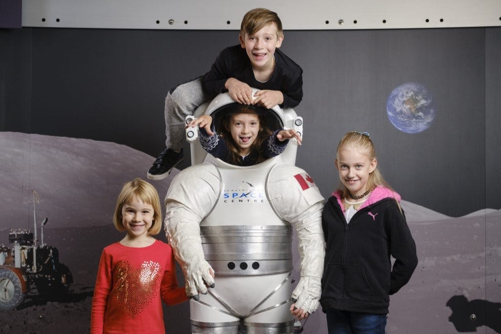 우주복 디스플레이를 들고 포즈를 취하는 네 명의 어린이; 한 명의 어린이가 양복 안에 있고 다른 한 명은 정상에 올라가고 있습니다. 달과 지구 배경.