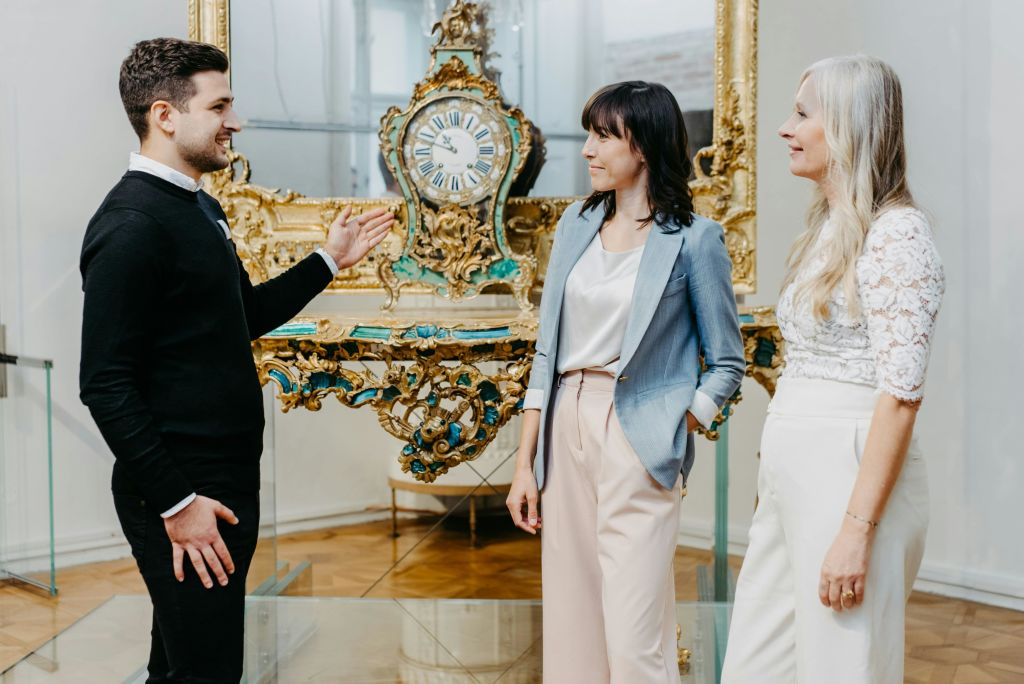 화려한 골동품 시계 근처에 두 명의 여성이 서 있으며, 세 번째 여성이 그 시계를 향해 손짓하고 있습니다. 장면이 실내에 있는 것 같습니다.