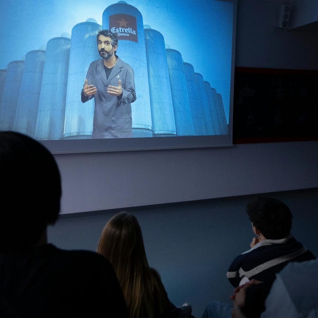 커다란 사일로를 배경으로 한 남자가 영사된 화면에서 말하는 것을 여러 사람이 지켜보고 있습니다.