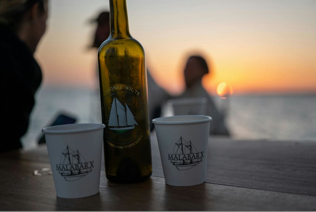 탁자 위에 선박 디자인이 있는 유리병과 종이컵 2개, 흐릿한 일몰과 배경의 사람들.