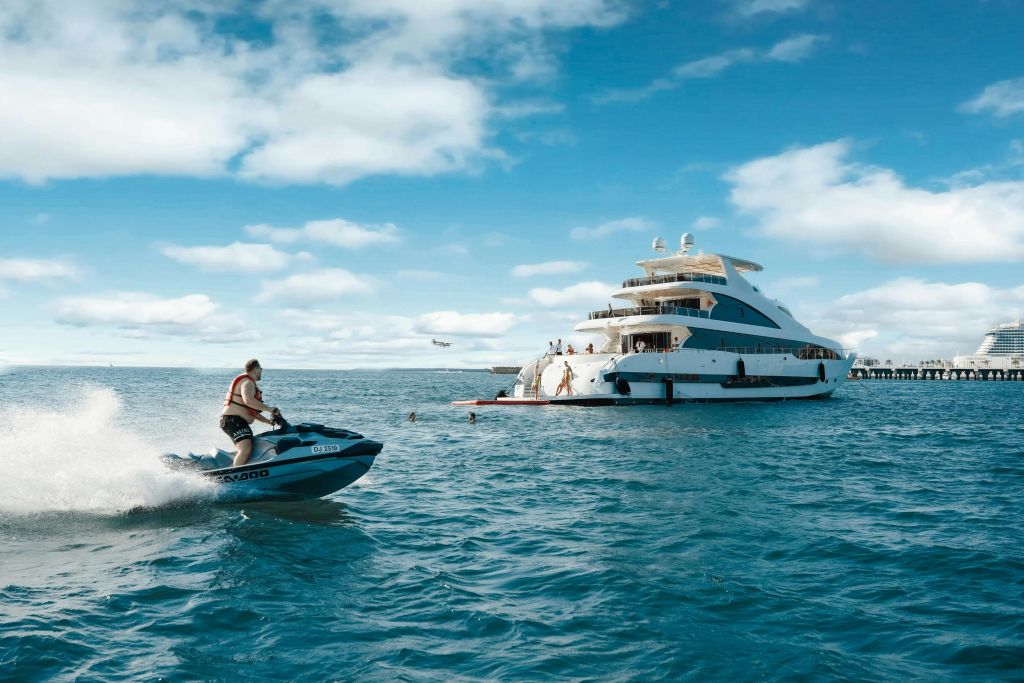 한 사람이 화창한 날 요트 갑판에서 다른 사람들과 함께 대형 요트 근처에서 제트 스키를 타고 있습니다.