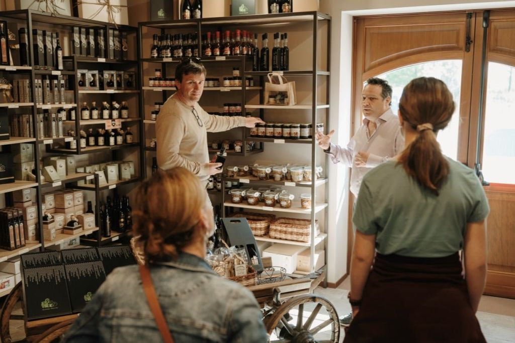 한 상점에서 4명이 항아리와 병이 놓인 선반 근처에 서 있습니다. 한 사람은 제품을 가리키고 다른 사람은 설명합니다.