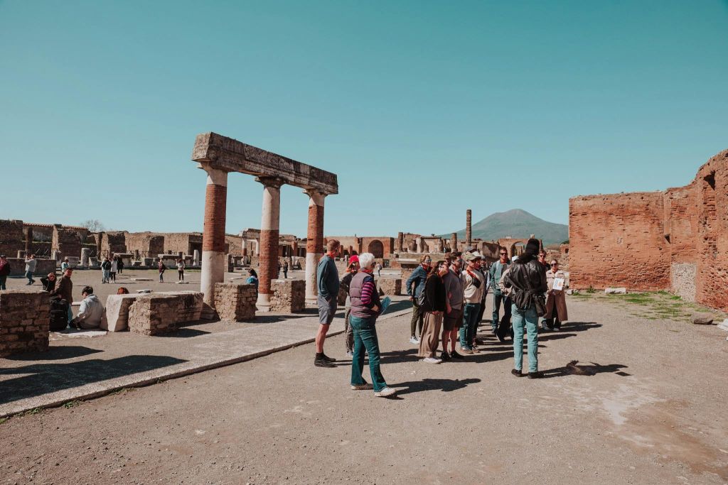 맑은 하늘 아래 기둥이 있고 먼 산을 배경으로 관광객들이 고대 유적 근처에 서 있습니다.