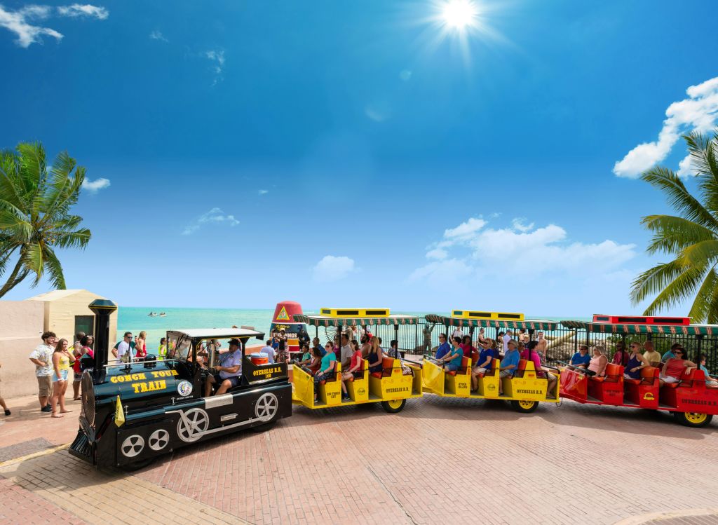 화창한 날 해변 근처에서 노란색 마차를 타고 푸른 하늘과 바다를 배경으로 작은 관광 열차를 타는 사람들.