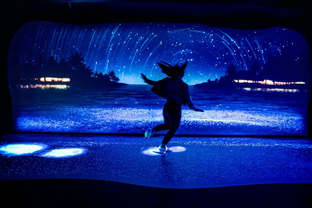 실루엣의 야자수와 빛나는 조명이 있는 해변 위로 별이 빛나는 밤하늘을 보여주는 디지털 화면 앞에서 한 사람이 춤을 추고 있습니다.
