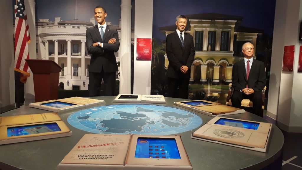 양복을 입은 남성의 왁스 피규어 두 개가 포즈를 취하고 있습니다. 하나는 커다란 건물을 배경으로 서서 앉고 하나는 지도와 문서가 있는 테이블입니다.