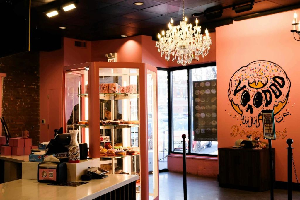 분홍색 벽, 페이스트리 진열장, 샹들리에, 창문 근처에 커다란 도넛을 테마로 한 벽화가 있는 아늑한 도넛 가게입니다.