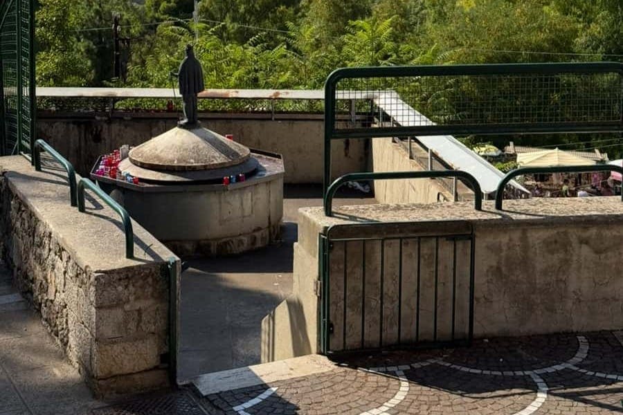 녹색과 울타리 통로로 둘러싸인 동상이 있는 작은 콘크리트 사당으로, 측면에는 빨간색과 파란색 촛불이 있습니다.