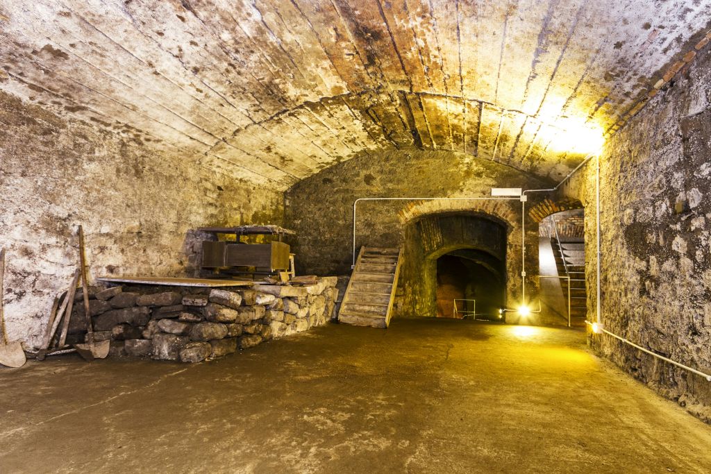 아치형 천장, 계단, 기계가 있는 높이 솟은 플랫폼, 희미한 조명이 있는 오래된 석조 저장고입니다.