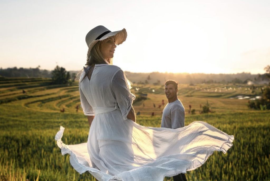 하얀 드레스와 모자를 쓴 여성이 해질녘 들판에 서 있고, 뒤에서 한 남성이 미소를 짓고 있습니다.