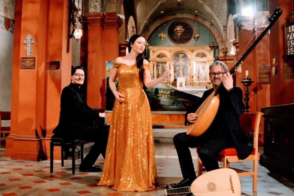 반짝이는 금색 드레스를 입은 한 여성이 화려하게 장식된 교회에서 피아니스트와 기타리스트 사이에서 노래를 부릅니다.